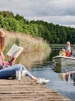 VRK Altersvorsorge – Eine junge Frau sitzt auf einem Steg an einem See während ihr Mann mit den Töchtern in einem Boot fährt.