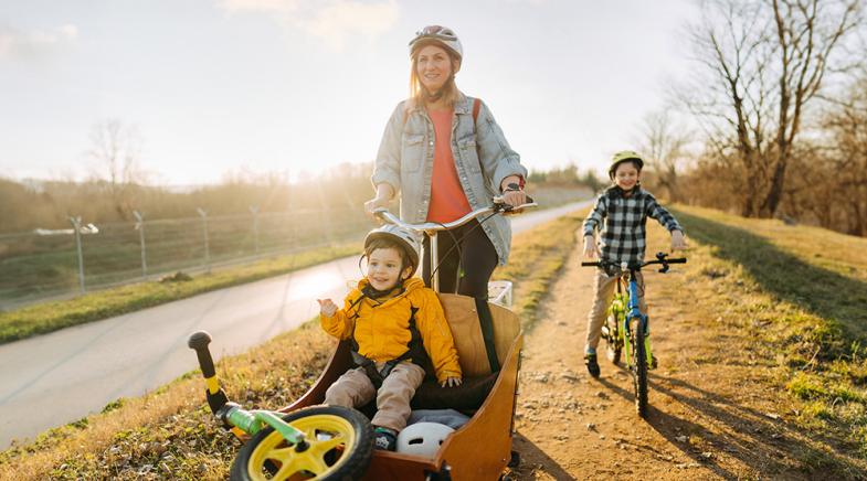Eine junge Frau fährt mit Ihren Kindern Fahrrad auf einem Radweg.